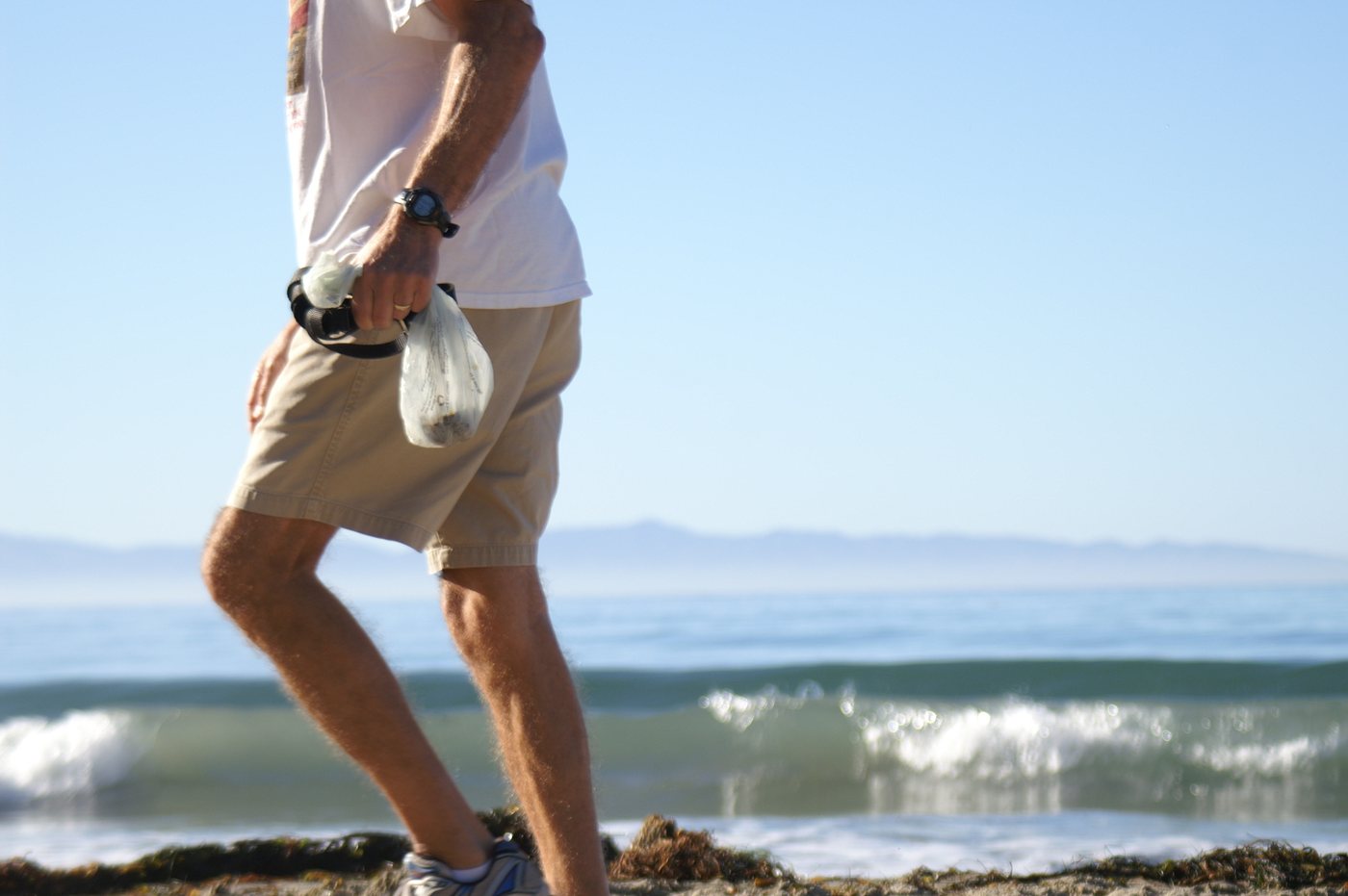 A man walks along the beach swinging a clear bag of dog poop from his hand, next to his leash